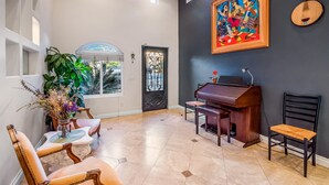 Een ruime kamer met een piano, een eettafel met stoelen, een groot raam en een deur met decoratieve ijzerwerk.