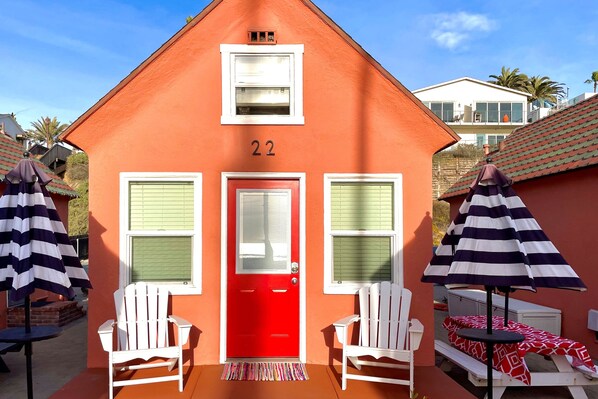 Une maison rouge portant le numéro 22, deux chaises blanches et des parasols à rayures.