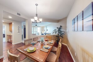 A dining area with a wooden table set for six, surrounded by chairs. There is a ceiling fan and a chandelier above the table.