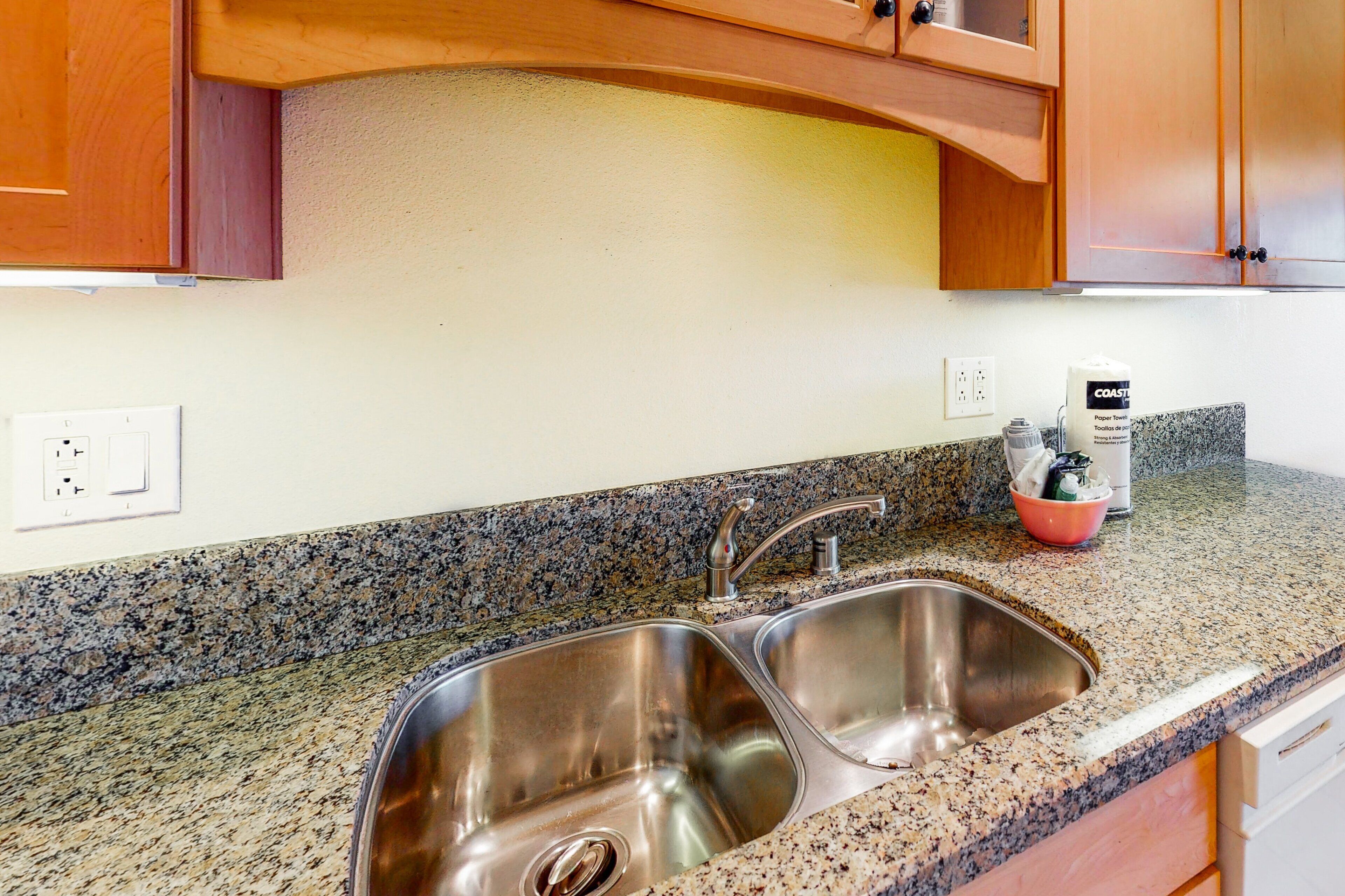 A kitchen with a double stainless steel sink, granite countertop, wooden cabinets, and a paper towel dispenser.