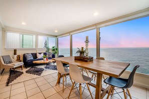 Amplia sala de estar con una gran mesa de madera, sillas blancas y un sofá. La habitación tiene ventanas amplias con vistas al océano y al atardecer.