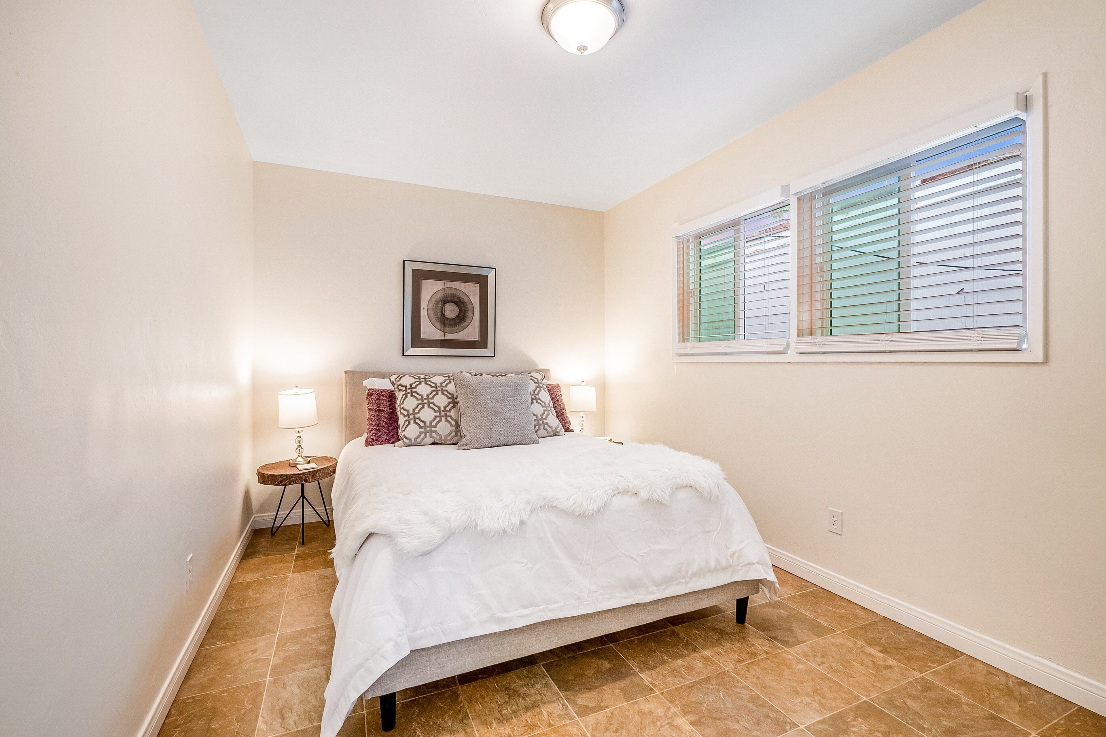 A bedroom with a bed, a bedside table with a lamp, a framed picture on the wall, and a window with blinds.