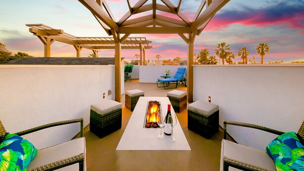 Una terrazza sul tetto con una pergola, poltroncine, un tavolo con un elemento decorativo a fuoco e vista su palme sullo sfondo del cielo al tramonto.