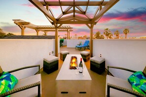 A rooftop terrace with a pergola, lounge chairs, a table with a fire feature, and a view of palm trees against a sunset sky. - New Tremont Modern Beach (Oceanside)