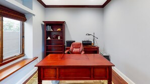 Ein Zimmer mit einem großen Holztisch, einem Ledersessel, einem Bücherregal und einem Fenster mit Jalousien.