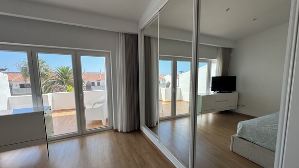 A modern bedroom with a large sliding glass door leading to a balcony, a flat-screen TV on a white dresser, and a bed with a grey bedspread.