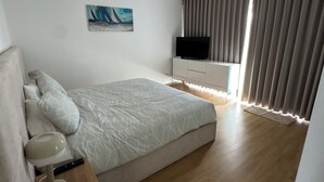A modern bedroom with a large bed, a bedside table with a lamp, a TV on a stand, and a painting on the wall.