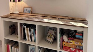 Una habitación con pared blanca, ventana con marcos blancos, un escritorio de madera y una estantería llena de libros y fotografías enmarcadas.