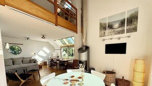 Un salón amplio con techo alto, una mesa de comedor con sillas blancas, una chimenea y un ventanal que ofrece luz natural.