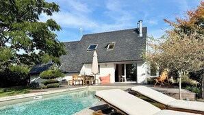 Una piscina con sillones de descanso, un patio con mobiliario exterior y una casa con tejado inclinado y paneles solares.