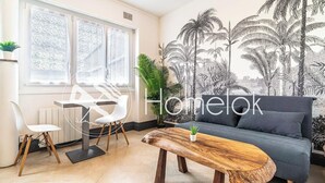 A modern living room with a grey sofa, a wooden coffee table, and a white chair. There is a large window with sheer curtains, a wall mural of palm trees, and a radiator. - Séjour en centre-ville - rénové et confortable (Lorient)