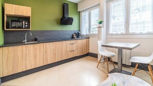 A modern kitchen with wooden cabinets, a black microwave, and a coffee machine. There is a window with a view of buildings outside. - Séjour en centre-ville - rénové et confortable (Lorient)
