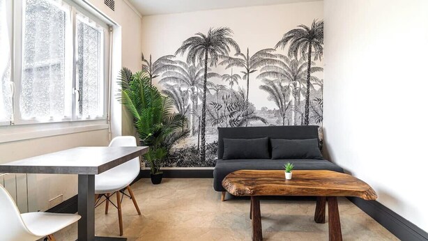 A modern living room with a large wall mural of palm trees, a grey sofa, a wooden coffee table, and a dining area with a white table and chairs. - Séjour en centre-ville - rénové et confortable (Lorient)