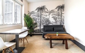 A modern living room with a large wall mural of palm trees, a grey sofa, a wooden coffee table, and a dining area with a white table and chairs. - Séjour en centre-ville - rénové et confortable (Lorient)