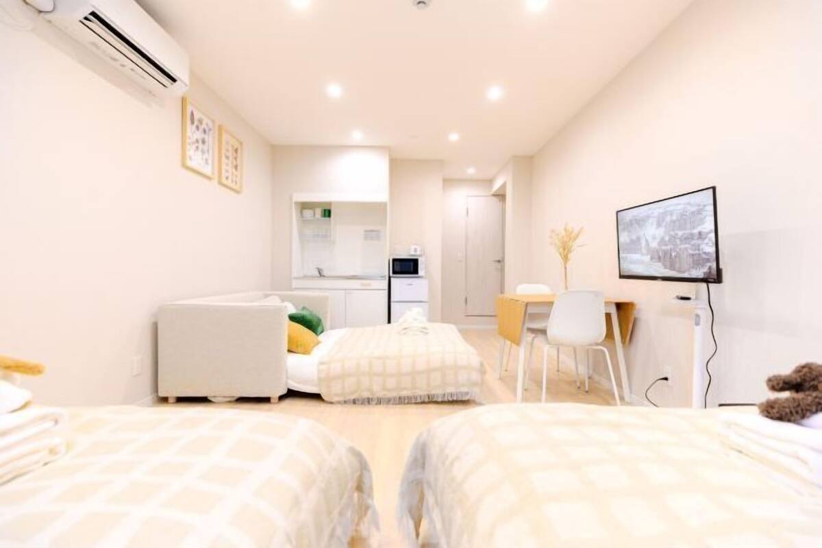 A modern hotel room with two beds, a desk, a chair, a TV, and a kitchenette in the background.