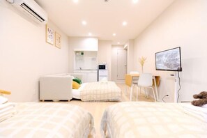 Une chambre d’hôtel moderne avec deux lits, un bureau, une chaise, un téléviseur et, au fond, une kitchenette.