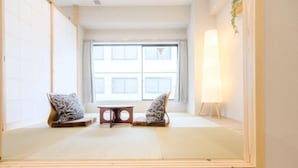 Una habitación con esterillas de tatami, una mesa baja y dos cojines, un ventanal grande y una lámpara fijada a la pared.