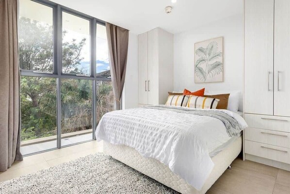 A bedroom with a large window, a bed with white bedding, and a view of greenery outside. - Comfy & Cozy Beach Studio 5min Walk to Bondi Beach (Sydney)
