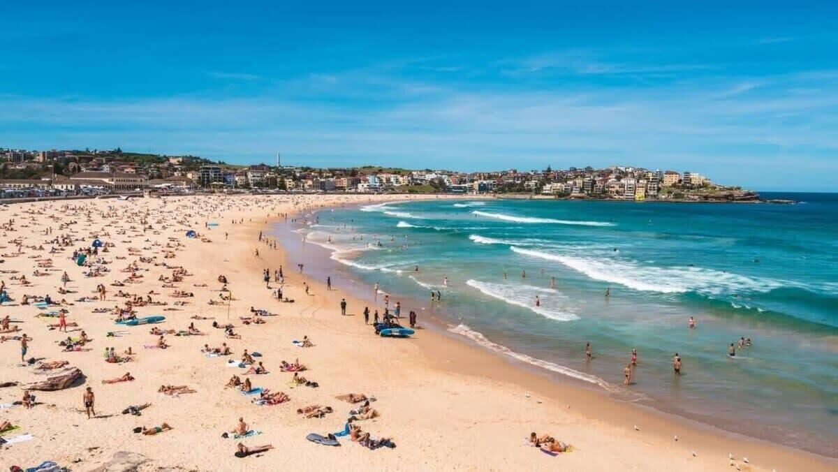 Ein überfüllter Strand mit badenden und sonnenden Menschen, ein klarer, blauer Himmel und Gebäude im Hintergrund.