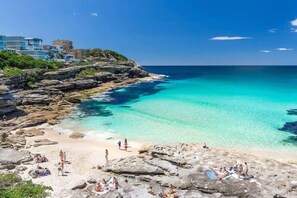 A coastal beach with clear turquoise waters, rocky shoreline, and buildings in the background. - Comfy & Cozy Beach Studio 5min Walk to Bondi Beach (Sydney)