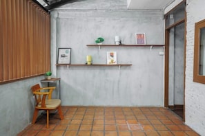 Una habitación pequeña con suelo de baldosas, una silla de madera, una estantería con libros y una planta, y una puerta.