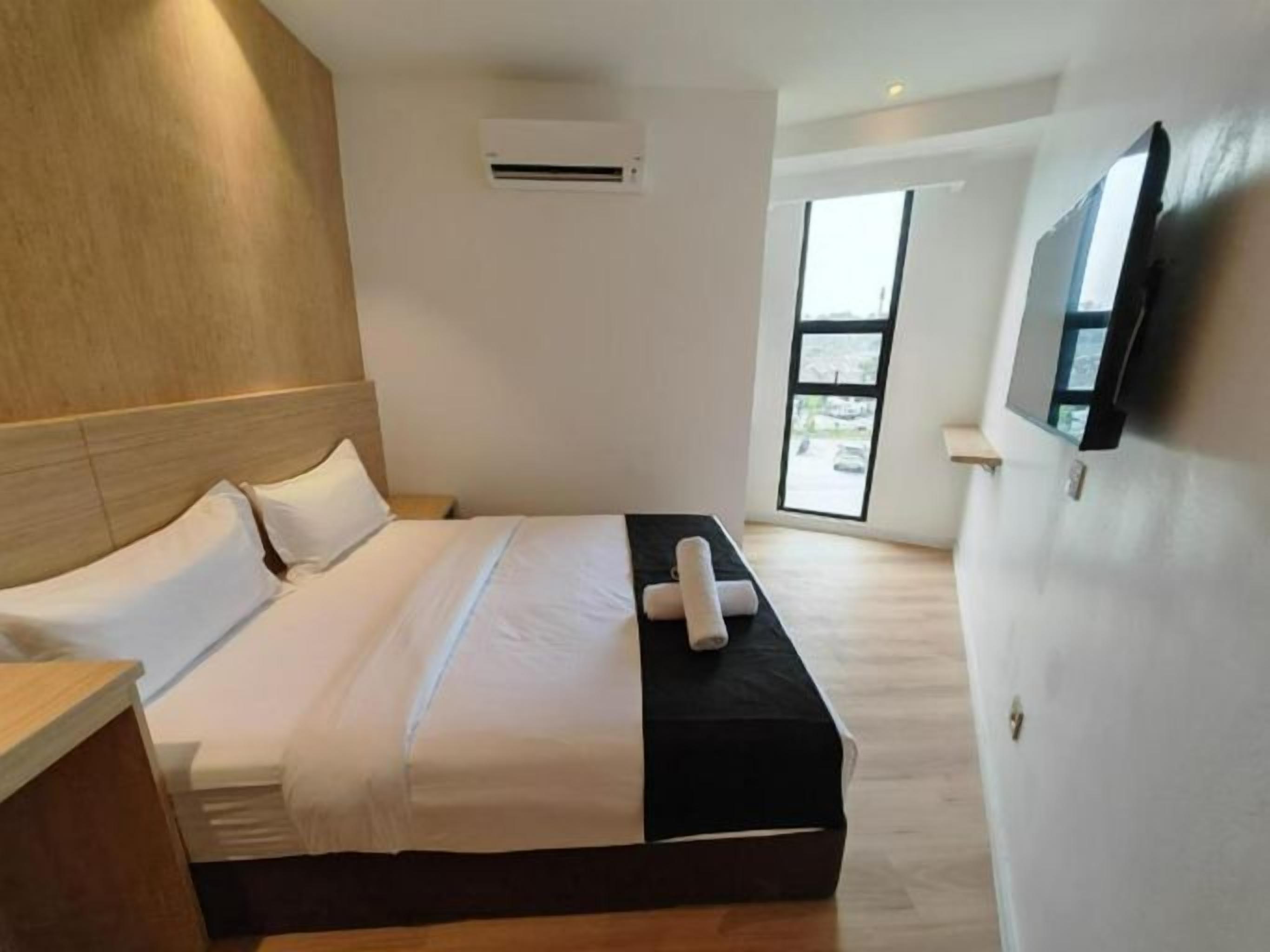 A modern hotel room with a large bed, a flat-screen TV mounted on the wall, and a window providing a view of the outdoors.