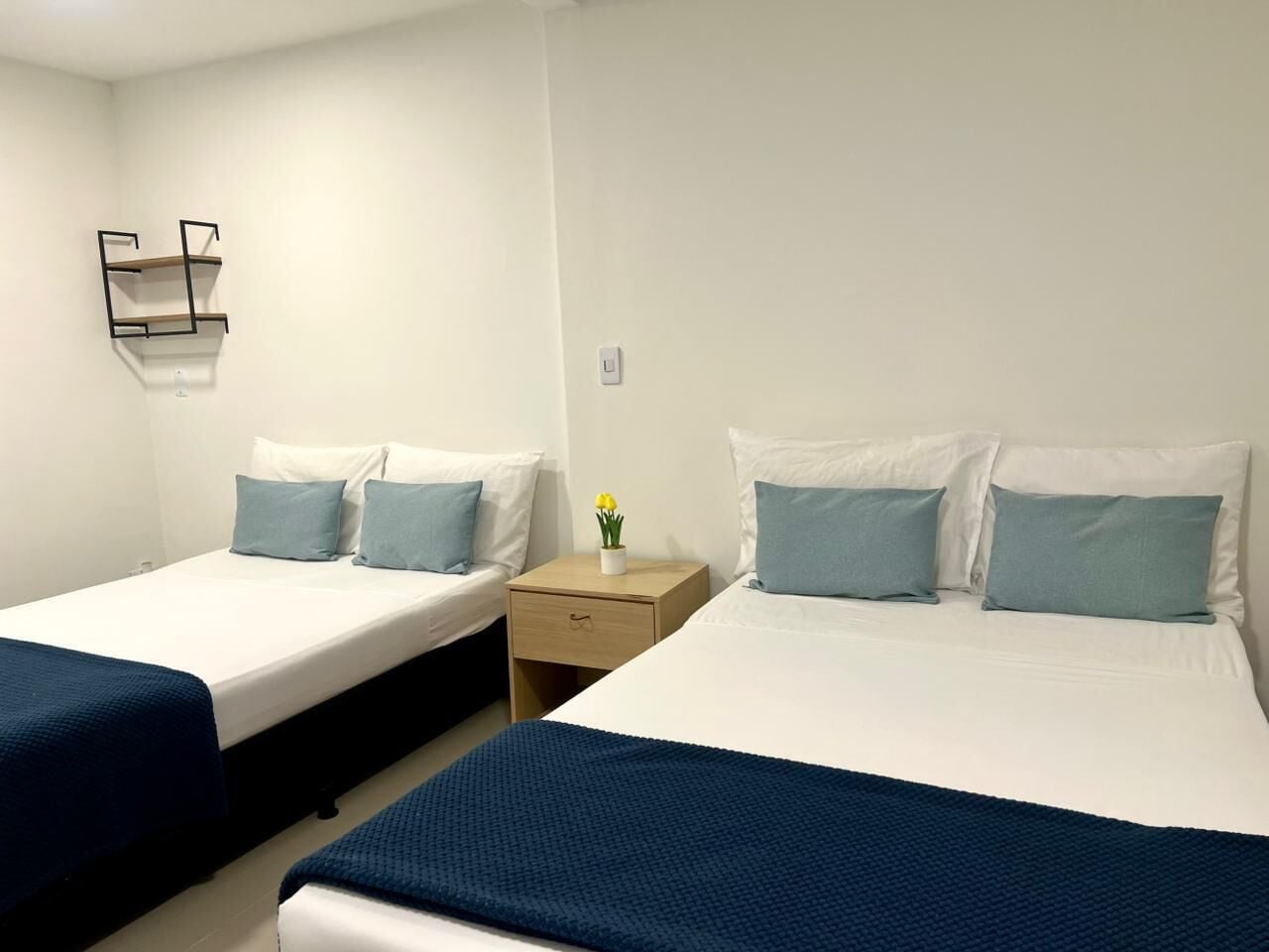 Een hotelkamer met twee éénpersoonsbedden, elk met een blauwe spreidek en passende kussens. Er staat een klein nachtkastje met een bloevas en een lade.