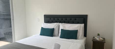 Una cama impecablemente arreglada con almohadas blancas y azul turquesa, una manta gris y una toalla blanca enrollada. Una mesita de noche de madera con una planta está al lado de la cama.