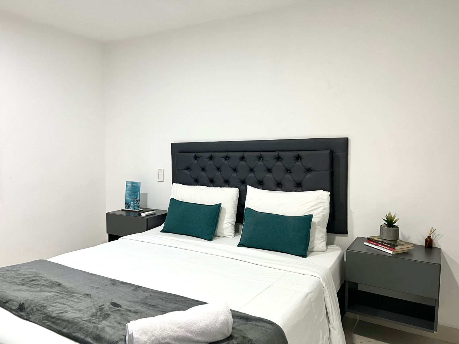 A neatly made bed with a black headboard, white bedding, and two teal pillows. There is a small bedside table with a book and a plant.