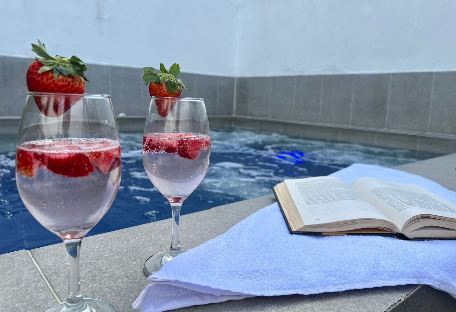 Zwei Gläser mit einem rosafarbenen Getränk, Erdbeeren, ein Buch und ein blaues Handtuch auf einer Steinoberfläche in der Nähe eines Pools.