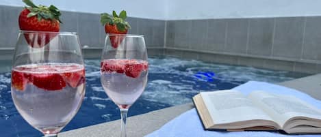 Twee glazen roze drank met aardbeien, een boek en een blauwe handdoek op een stenen oppervlak in de buurt van een zwembad.