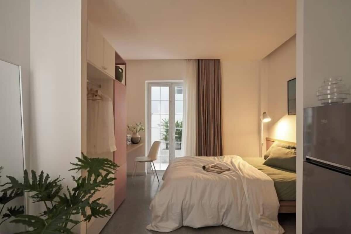 A bedroom with a bed, a chair, a desk, a plant, and a window with curtains.