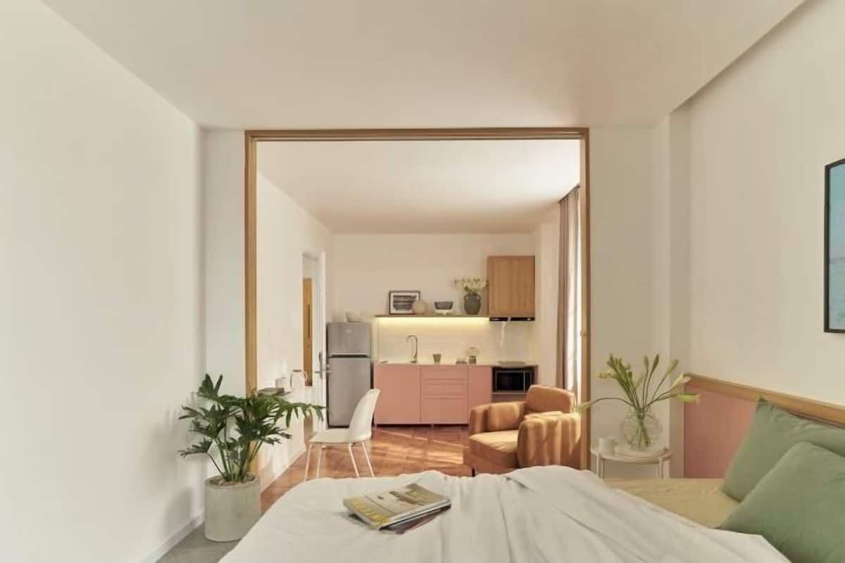 A compact hotel room with a bed, bedside table, and a view into the kitchen area.