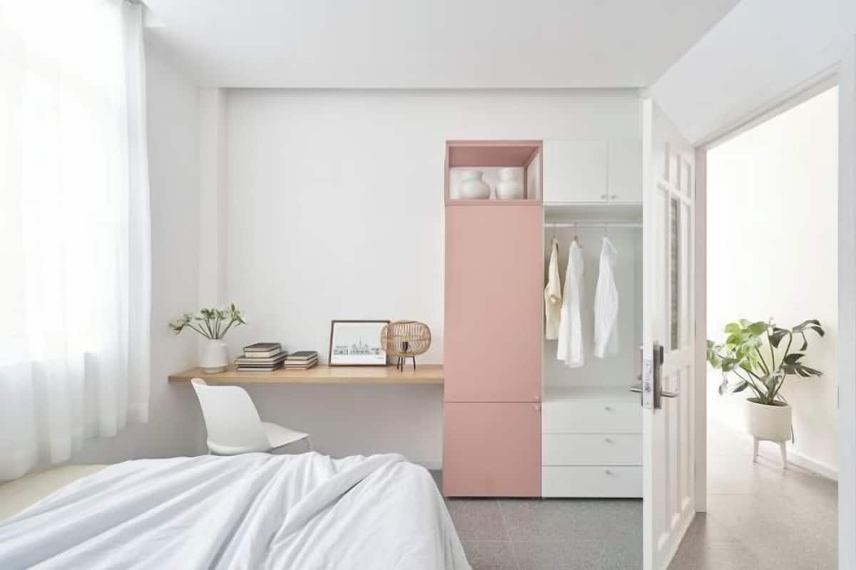 A bedroom with a bed, a desk with a pink cabinet, a wardrobe, and a potted plant.