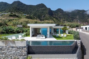 A modern house with a swimming pool, a patio with lounge chairs, and a view of a mountainous landscape. - Villa Guindaste I – Modern Villa with Sea View (Faial)
