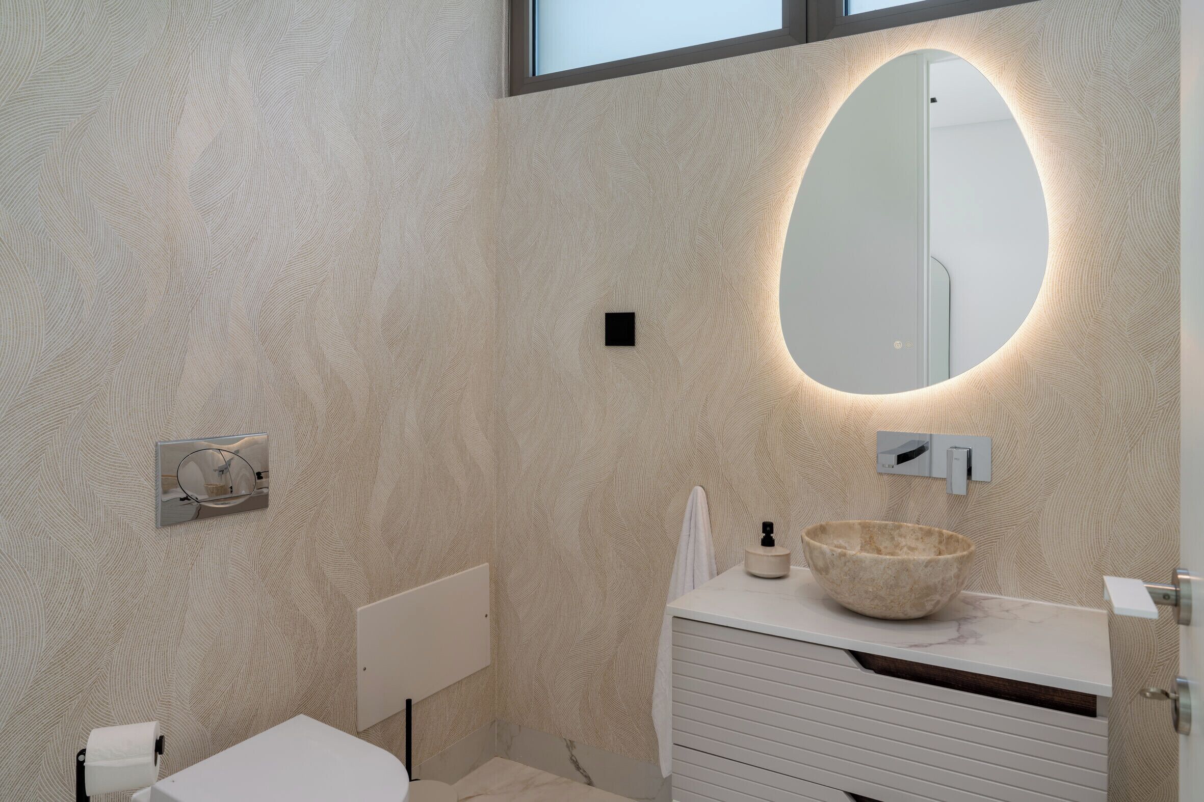 A modern bathroom with a textured wall, a round mirror, a white sink with a stone basin, and a toilet.