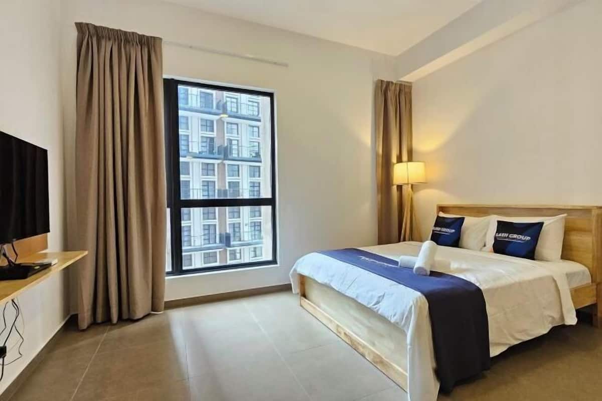 A hotel room with a large window, a bed with a blue headboard, a bedside lamp, and a television on a wooden stand.