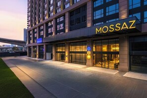 Un hotel moderno con un llamativo letrero 'MOSSAZ', un acceso cubierto y un área de estacionamiento.