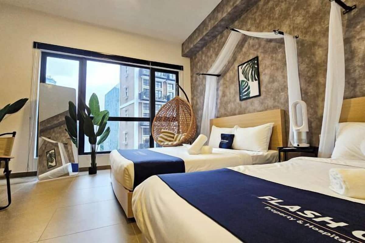 A hotel room with two beds, a large window, a hanging chair, and a view of buildings outside.
