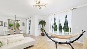 Un amplio espacio de sala con una hamaca, un sofá blanco, una mesa de comedor con sillas y un gran ventanal con cortinas.