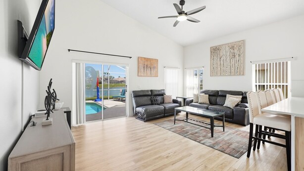 A spacious living room with a flat-screen TV, a ceiling fan, and a pool view through glass doors.
