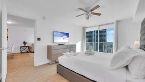 Une chambre moderne avec un lit, un ventilateur de plafond, une télévision et une vue sur le paysage urbain, visible à travers de grandes fenêtres.