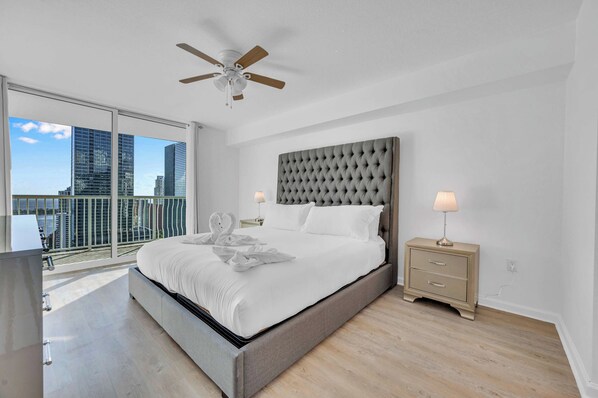 A modern bedroom with a large bed, a ceiling fan, a nightstand with a lamp, and a view of the city through a balcony.