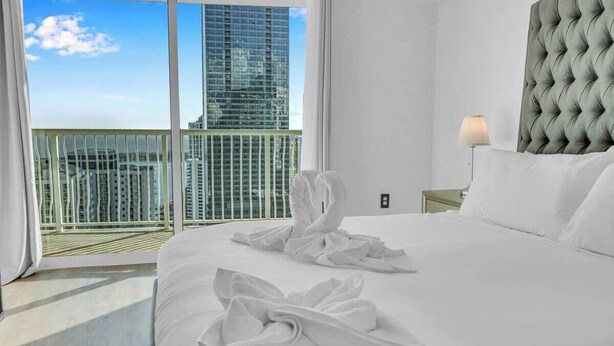 Une chambre d’hôtel avec un grand lit, une tête de lit capitonnée, une table de chevet avec une lampe et une vue sur le paysage urbain depuis un balcon.