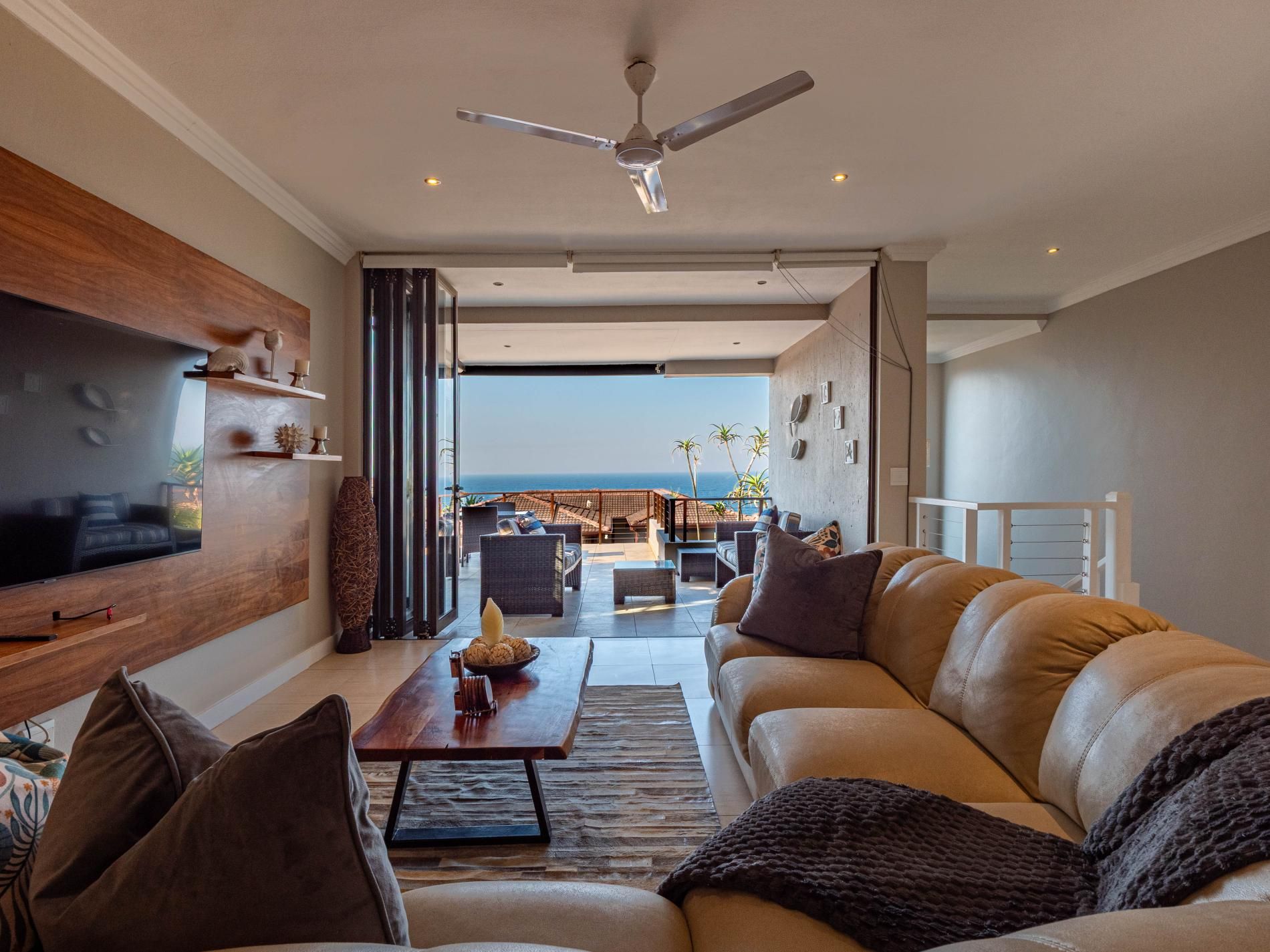 A modern living room with a leather sofa, a flat-screen TV mounted on a wooden wall, and a ceiling fan.