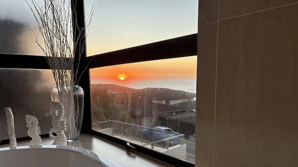 Ein Badezimmer mit einer Badewanne, einer Vase mit Zweigen und Blick auf einen Sonnenuntergang über einer Stadtlandschaft.
