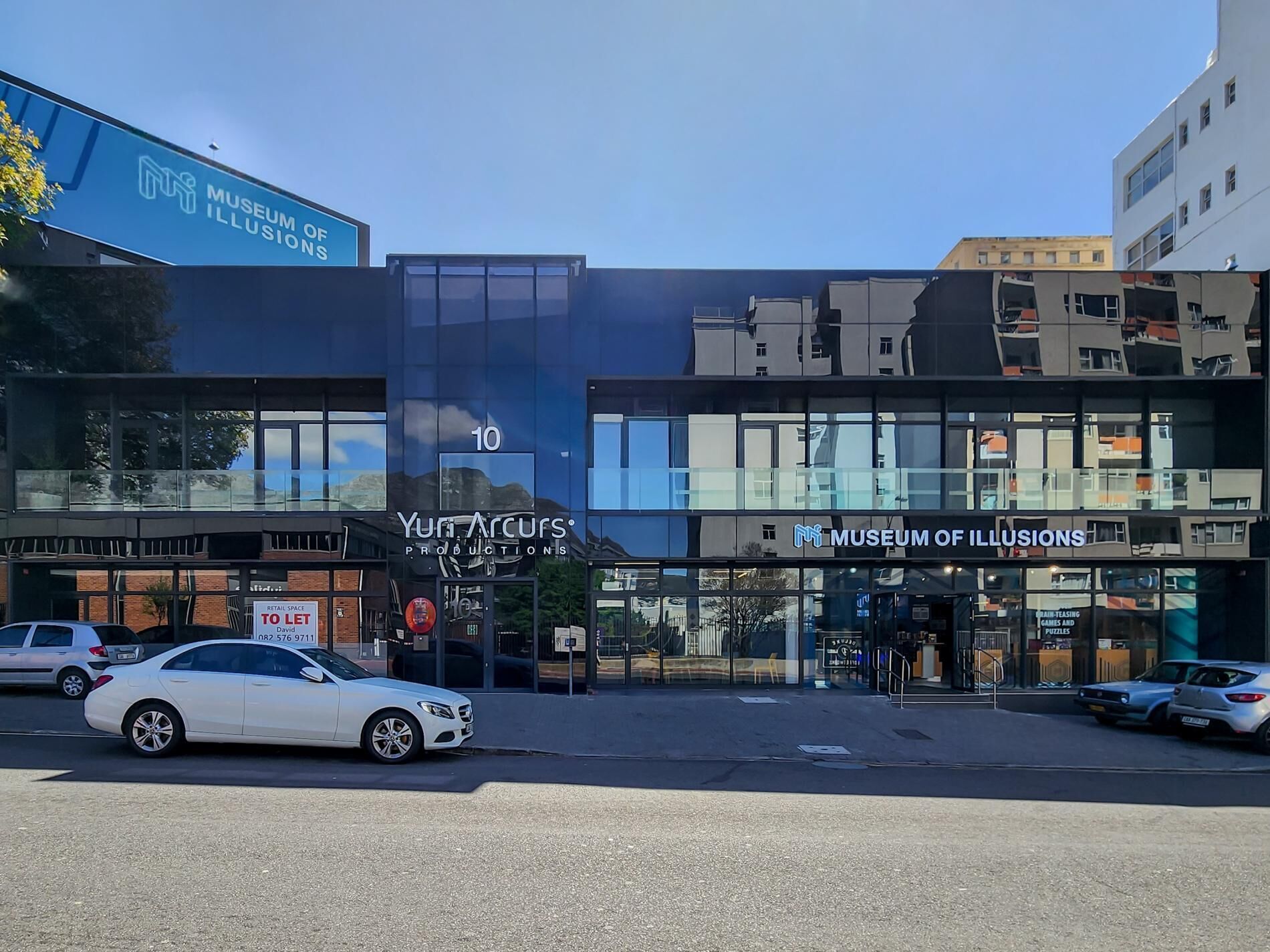 Un bâtiment moderne à la façade vitrée, sur lequel on peut voir les enseignes « Musée des illusions » et « Yumi Arcuris Productions ».