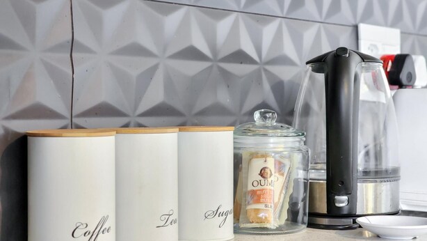 Un plan de travail de cuisine avec trois récipients blancs étiquetés « Café », « Thé » et « Sucre », un bocal en verre avec une étiquette, une machine à café noire et un petit bol blanc.
