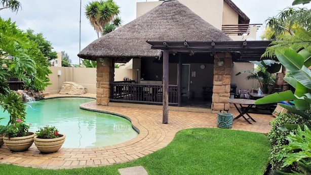 A backyard with a swimming pool, a thatched-roof gazebo, and a patio area with outdoor furniture.