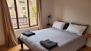 A neatly made bed with a grey bedspread and two pillows, a bedside lamp, and a view of a balcony with a palm tree.